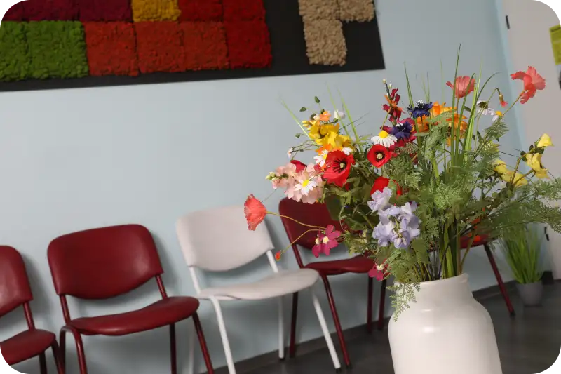 Das Bild zeigt den Wartebereich der Hausarztpraxis in Hormersdorf im Erzgebirge, gestaltet mit modernen roten und weißen Stühlen und einem kleinen Tisch, auf dem ein farbenfroher Blumenstrauß steht. Die freundliche Einrichtung vermittelt eine angenehme und beruhigende Atmosphäre, die Patienten bereits beim Eintreten willkommen heißt. 